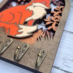 May include: Wooden key holder with a fox silhouette in an archway design. The fox is orange and white and is surrounded by brown leaves. There are three brass hooks on the bottom.