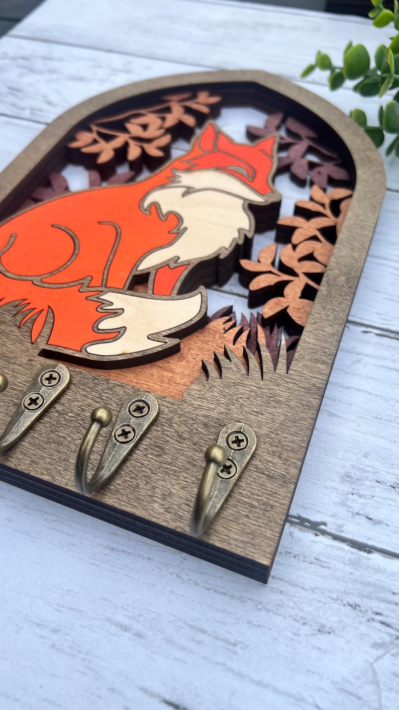 May include: Wooden key holder with a fox silhouette in an archway design. The fox is orange and white and is surrounded by brown leaves. There are three brass hooks on the bottom.