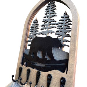 May include: Wooden key holder with a bear and pine tree silhouette design. The key holder has a natural wood finish and features five black metal hooks for hanging keys. The design is cut out of a dark brown material.
