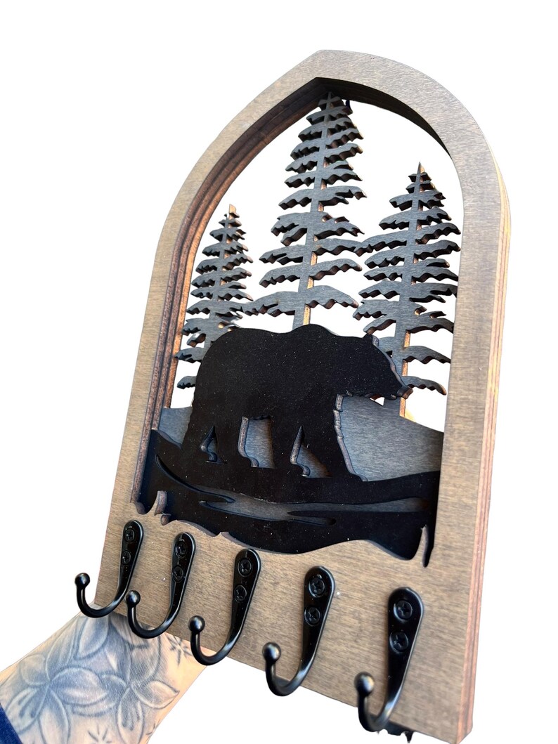 May include: Wooden key holder with a bear and pine tree silhouette design. The key holder has a natural wood finish and features five black metal hooks for hanging keys. The design is cut out of a dark brown material.