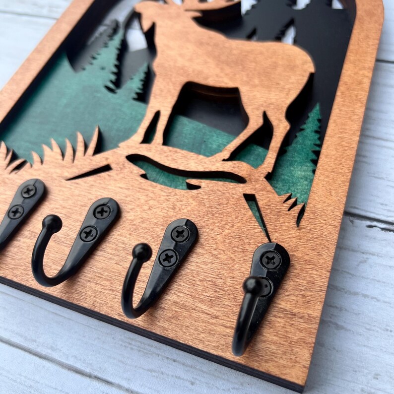 May include: Wooden key holder with a cutout of a moose standing in a forest scene. The key holder has four black metal hooks.