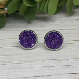 May include: A pair of small, round stud earrings with a silver setting and a purple glitter center.