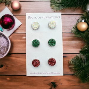 May include: A set of six round stud earrings in three colors: iridescent white, green, and red. Each earring has a glittery, textured surface and a silver-colored metal border. The card reads "Bigfoot Creations" and "Handmade in Northern Minnesota."