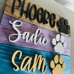 Puede incluir: Un letrero de madera con tres nombres, "Phoebe", "Sadie" y "Sam", pintados en diferentes colores. Cada nombre est&aacute; acompa&ntilde;ado de una huella de pata de un color diferente: negro, blanco y dorado.