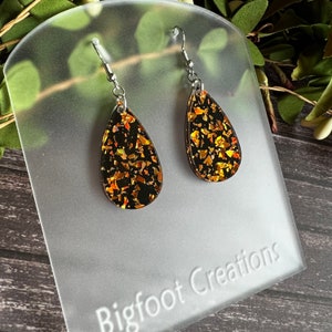 May include: A pair of teardrop-shaped earrings with black resin and gold glitter. The earrings are on a white background with the text "Bigfoot Creations".