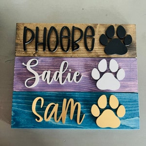 Puede incluir: Tres letreros de madera con los nombres "Phoebe", "Sadie" y "Sam" escritos en letras negras, blancas y doradas, respectivamente. Cada letrero presenta una huella de pata de un color diferente: negro, blanco y dorado.