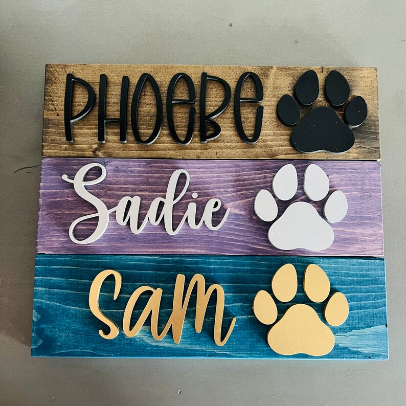 Puede incluir: Tres letreros de madera con los nombres "Phoebe", "Sadie" y "Sam" escritos en letras negras, blancas y doradas, respectivamente. Cada letrero presenta una huella de pata de un color diferente: negro, blanco y dorado.