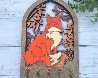 May include: Wooden key holder with a fox silhouette in orange and brown, surrounded by leafy branches. The key holder has five hooks.