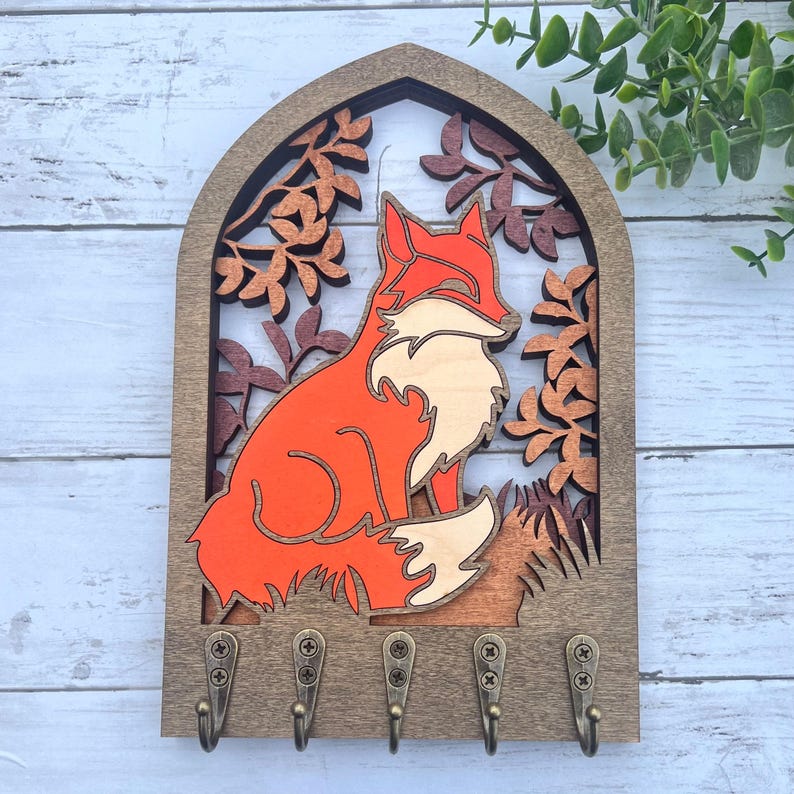 May include: Wooden key holder with a fox silhouette in orange and brown, surrounded by leafy branches. The key holder has five hooks.