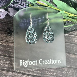 May include: A pair of teardrop-shaped earrings with a floral pattern in shades of green and white. The earrings are on a clear display stand with the text "Bigfoot Creations".