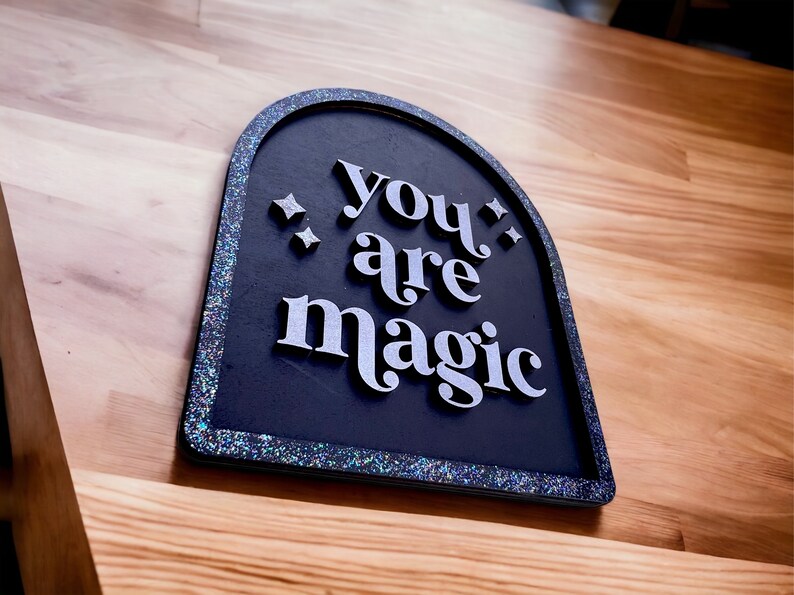 Pu&ograve; includere: Un cartello nero con un bordo scintillante recita "you are magic" in lettere bianche. Il cartello ha la forma di un arco.