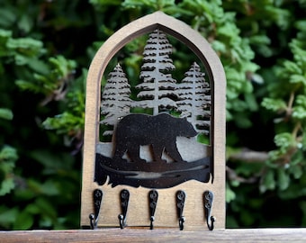 May include: Wooden key holder featuring a bear silhouette walking in front of a forest scene. The key holder has a dark brown finish and includes five black metal hooks for hanging keys. The design is framed by an arched top.