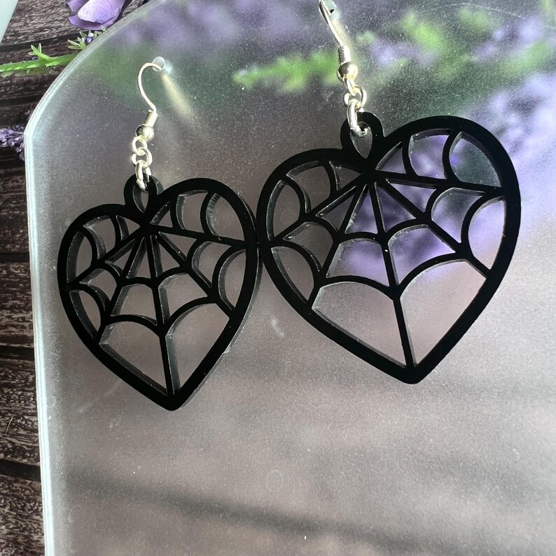 May include: A pair of black heart-shaped earrings with a spiderweb design. The earrings are made of acrylic and have silver hooks.
