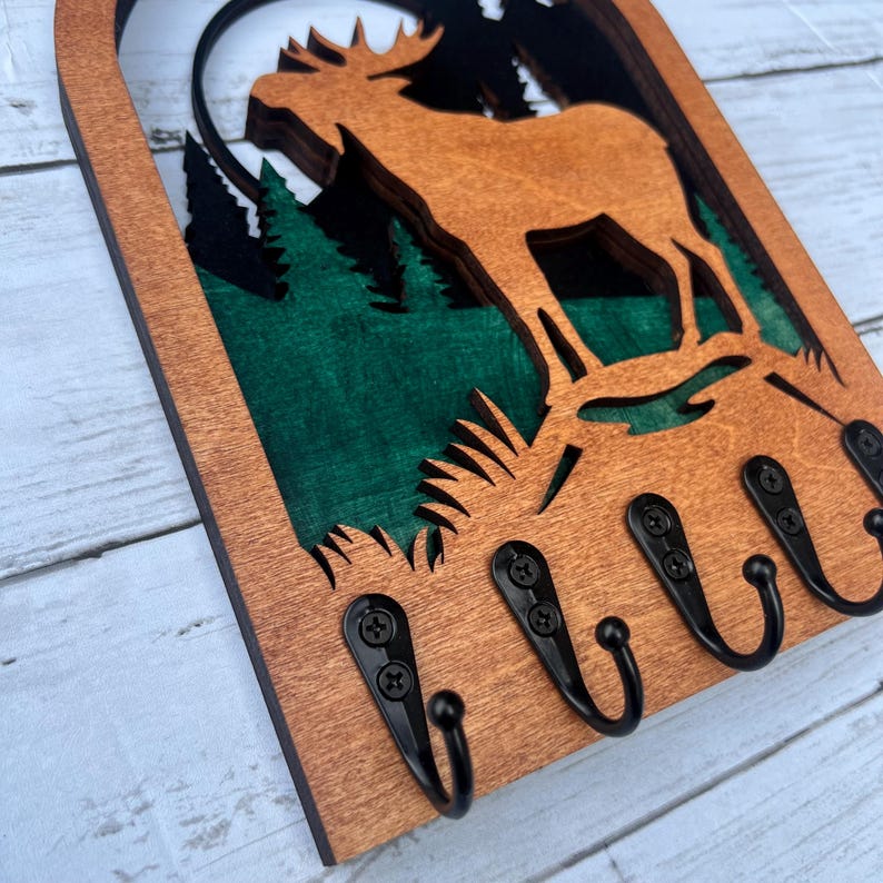 May include: Wooden key holder with a moose silhouette and a green forest background. The key holder has five black metal hooks.