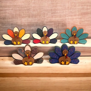 May include: Six wooden turkey figurines with different colored and patterned feathers. The turkeys are all facing forward with their heads tilted slightly down. The turkeys are all standing on a wooden surface.