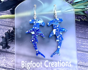 May include: A pair of blue hammerhead shark earrings with a white and blue patterned design. The earrings are on a white display card with the text "Bigfoot Creations".