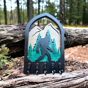 May include: A black wooden key holder shaped like an archway with a silhouette of Bigfoot walking through a forest. The forest is painted in shades of green and brown. There are five hooks on the bottom of the key holder.