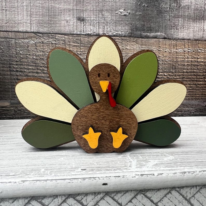 May include: A wooden turkey figurine with a brown body, green and white feathers, and a red wattle. The turkey has yellow feet and is sitting on a white surface.