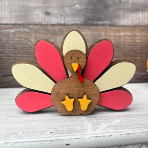 May include: A wooden turkey figurine with a brown body, red and white feathers, and yellow feet. The turkey is standing on a white surface.