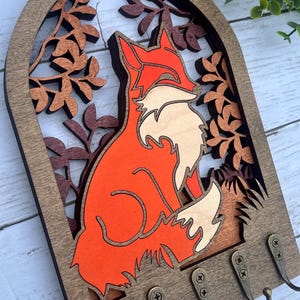 May include: Wooden key holder with a fox silhouette in orange and white, surrounded by a leafy design. The key holder has five hooks.