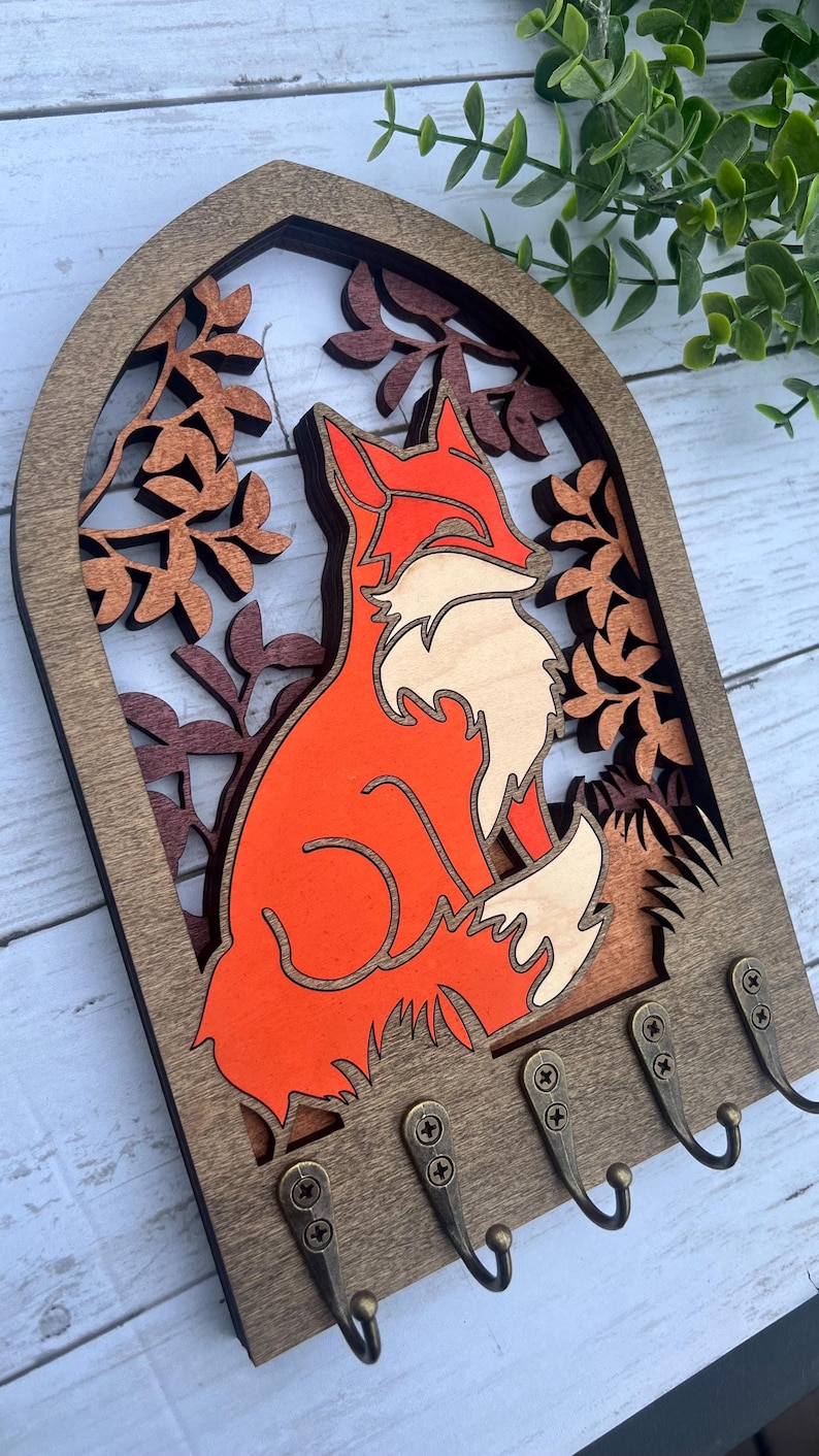 May include: Wooden key holder with a fox silhouette in orange and white, surrounded by a leafy design. The key holder has five hooks.