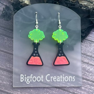 May include: A pair of earrings featuring a black beaker with a pink liquid and a green cloud on top. The earrings are on a white card with the text "Bigfoot Creations".