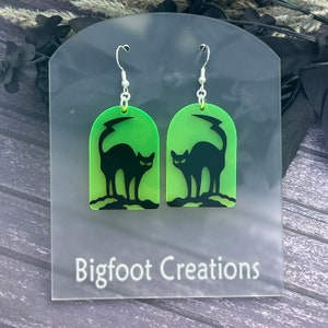 Puede incluir: Un par de pendientes de color verde y negro con silueta de gato. Los pendientes tienen forma de arco y presentan una silueta de gato negro con una cola de rayo. Los pendientes están en una tarjeta blanca con el texto "Bigfoot Creations".