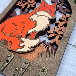May include: Wooden key holder with a fox silhouette in orange and white, surrounded by layered wood cutouts of leaves and branches. The key holder has three hooks for hanging keys.