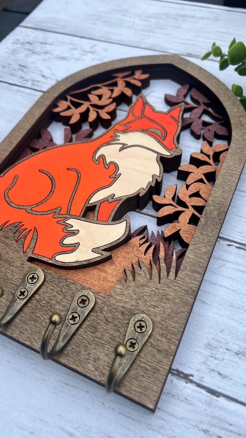 May include: Wooden key holder with a fox silhouette in orange and white, surrounded by layered wood cutouts of leaves and branches. The key holder has three hooks for hanging keys.