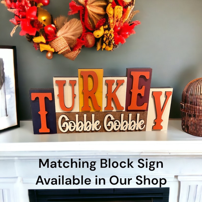 May include: A wooden block sign spelling "TURKEY" with the words "Gobble Gobble" underneath. The sign is decorated with fall colors and is displayed on a white mantel.