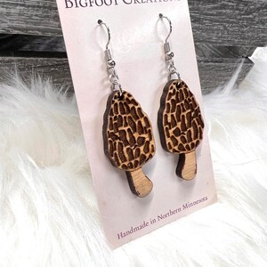 May include: A pair of wooden earrings shaped like morel mushrooms. The earrings are brown and have a natural wood finish. The earrings are hanging from silver hooks. The card reads "BIGFOOT CREATIONS" and "Handmade in Northern Minnesota".