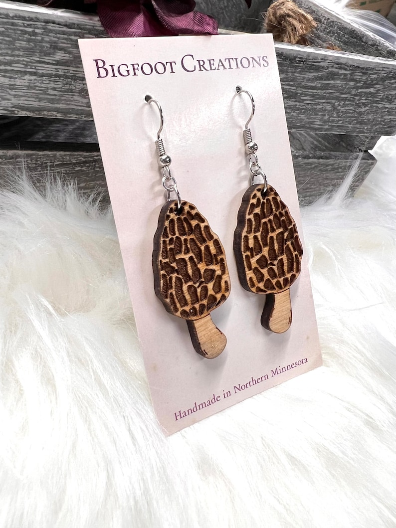 May include: A pair of wooden earrings shaped like morel mushrooms. The earrings are brown and have a natural wood finish. The earrings are hanging from silver hooks. The card reads "BIGFOOT CREATIONS" and "Handmade in Northern Minnesota".