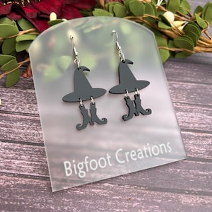 May include: Black witch earrings with a witch hat and boots silhouette. The earrings are on a white card with the text "Bigfoot Creations".