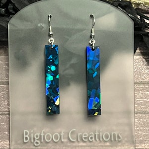 Puede incluir: Un par de pendientes rectangulares colgantes de color azul y verde iridiscente con ganchos plateados. Los pendientes se muestran en una tarjeta blanca con el texto "Bigfoot Creations".