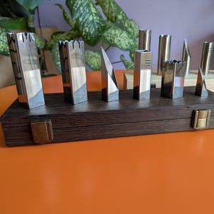 Op de afbeelding: Een verzameling metalen sculpturen in verschillende geometrische vormen, gepresenteerd op een donkere houten kist met messingkleurige sluitingen. De sculpturen hebben een zilver- en goudkleur, sommige met een kasteelachtig ontwerp.