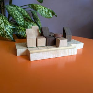 May include: A wooden architectural model with various geometric shapes, including cubes, rectangles, and triangular prisms. The model is constructed from light and dark wood tones, arranged on a rectangular base. The background features a plant with green and yellow leaves.