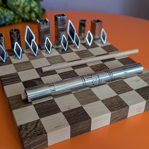May include: A unique chess set with metal pieces and a wooden board. The chess pieces include diamond-shaped and rectangular designs. A silver pen rests on the board, which has a checkerboard pattern. The board is approximately 12 inches square.