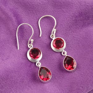 Puede incluir: Un par de pendientes colgantes de plata con piedras preciosas rojas vibrantes. Cada pendiente presenta una piedra redonda sobre una piedra en forma de lágrima, todo engastado en un bisel de plata. Los pendientes cuelgan de un gancho de plata simple.