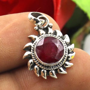 Pode incluir: Um pingente de prata com uma gema de rubi vermelho engastada em um design de sol.