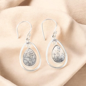 May include: A pair of silver dangle earrings. Each earring features a teardrop-shaped stone with a white and gray pattern, set within a silver teardrop frame. The earrings hang from a simple silver hook.