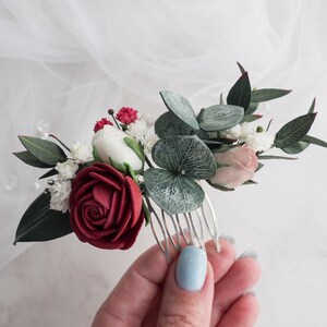 May include: A floral hair comb featuring a burgundy rose, white and pink flowers, and green leaves. The comb is made of silver metal and is adorned with small red buds and green eucalyptus leaves. This hair accessory is perfect for weddings or special occasions.