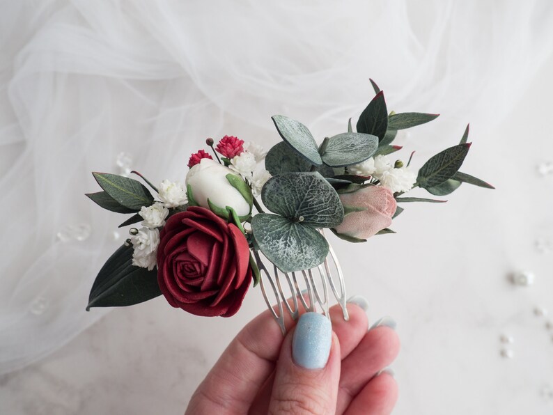 May include: A floral hair comb featuring a burgundy rose, white and pink flowers, and green leaves. The comb is made of silver metal and is adorned with small red buds and green eucalyptus leaves. This hair accessory is perfect for weddings or special occasions.