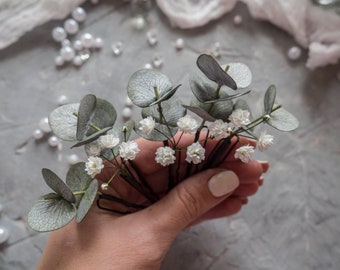 May include: A set of hair clips with small white flowers and eucalyptus leaves. The clips are held in a hand.