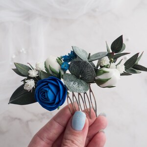 May include: A floral hair comb featuring a vibrant blue rose, white buds, and delicate white flowers. Green leaves and blue accents complement the design. The comb is ideal for weddings or special occasions.