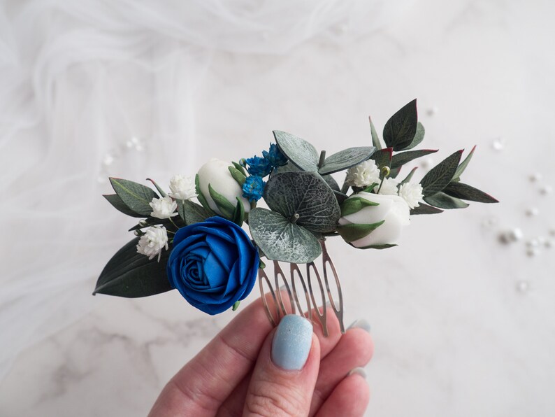 May include: A floral hair comb featuring a vibrant blue rose, white buds, and delicate white flowers. Green leaves and blue accents complement the design. The comb is ideal for weddings or special occasions.