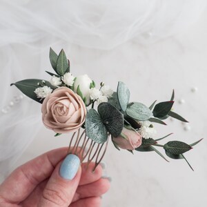 May include: A floral hair comb featuring a blush rose, white flowers, and green leaves. The comb is made of metal and is adorned with delicate, handcrafted flowers, perfect for weddings or special occasions.