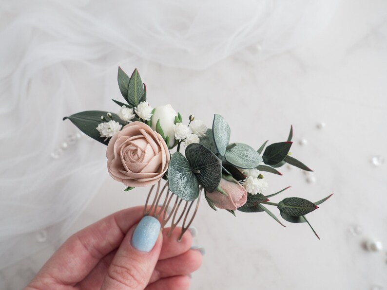 May include: A floral hair comb featuring a blush rose, white flowers, and green leaves. The comb is made of metal and is adorned with delicate, handcrafted flowers, perfect for weddings or special occasions.