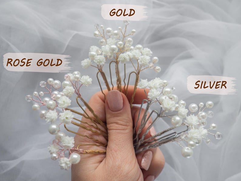 May include: A collection of hair pins featuring white floral accents, small clear beads, and white pearls on wire stems. The pins are available in three metal finishes labeled as GOLD, ROSE GOLD, and SILVER, each attached to a metal hair pin base.