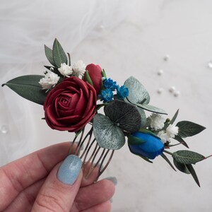 May include: A floral hair comb featuring a burgundy rose, blue and white accents, and green leaves. The comb is designed with a variety of artificial flowers and foliage, perfect for weddings or special occasions.