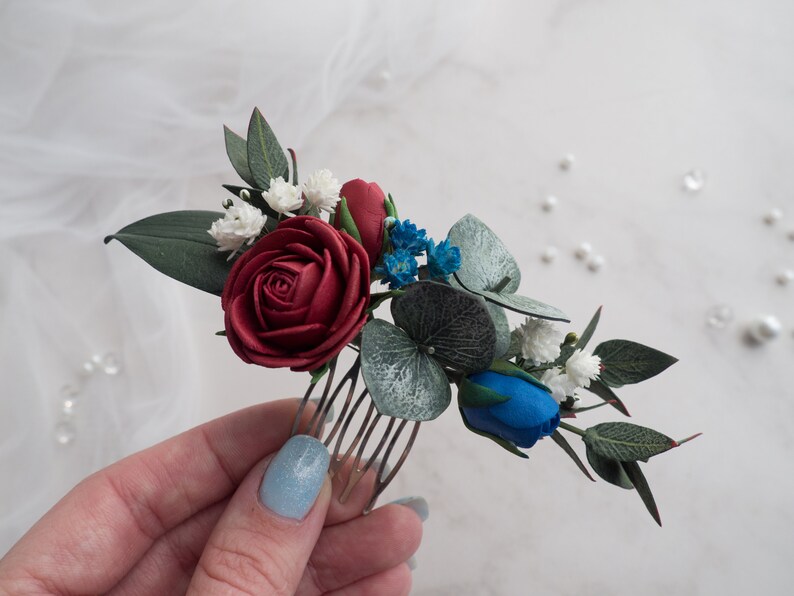 May include: A floral hair comb featuring a burgundy rose, blue and white accents, and green leaves. The comb is designed with a variety of artificial flowers and foliage, perfect for weddings or special occasions.